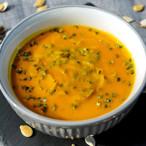 A bowl of vibrant orange Indian pumpkin soup, garnished with chopped green herbs and pumpkin seeds.