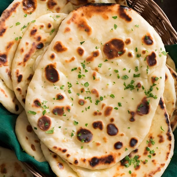Close-up of several pieces of golden-brown naan bread, garnished with fresh green herbs, in a basket.