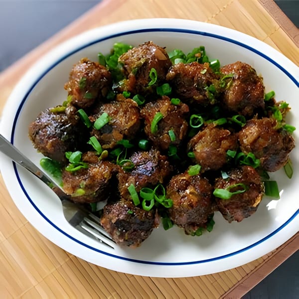 A white oval plate with blue trim holds brown Indian meatballs garnished with green onions and a fork.