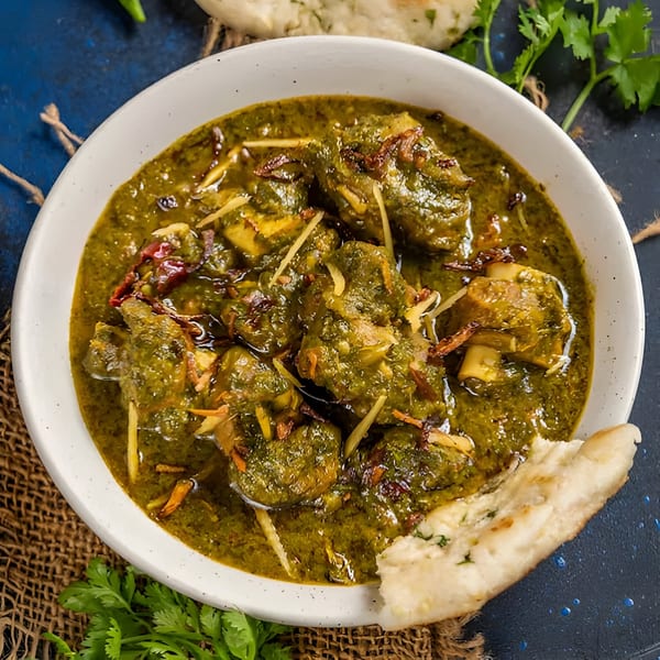A white bowl of green Indian curry with meat, garnished with ginger and fried onions, with naan bread.